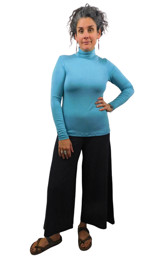 A woman modeling a bright turquoise blue long-sleeve mock neck turtleneck top with black wide-leg pants and brown sandals, standing against a plain white background.