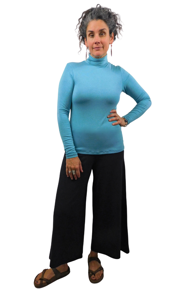 A woman modeling a bright turquoise blue long-sleeve mock neck turtleneck top with black wide-leg pants and brown sandals, standing against a plain white background.
