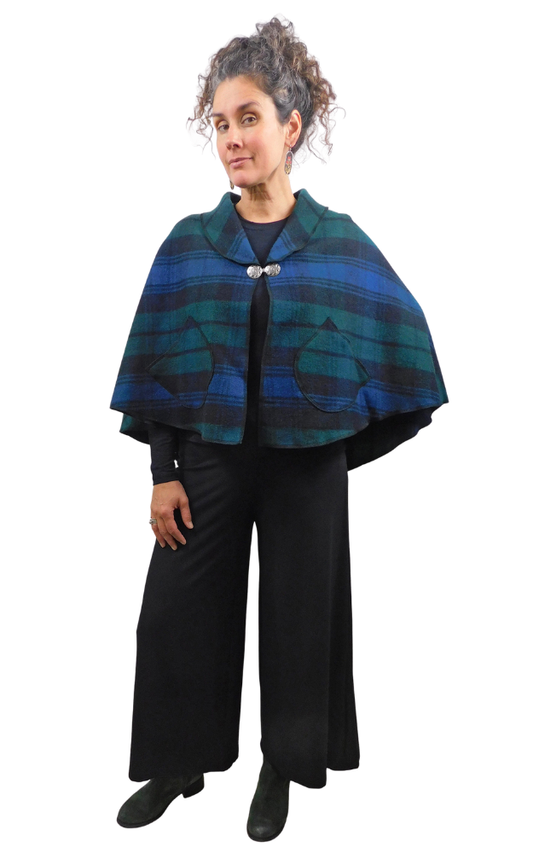 A woman modeling a blue and green plaid cape with a silver button closure, worn over a black outfit against a plain white background.