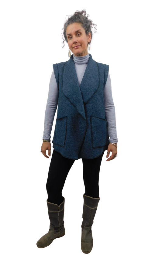 Woman wearing a dark blue heavy wool vest with large patch pockets, wearing a long sleeve turtleneck grey sweater and black tights and grey boots