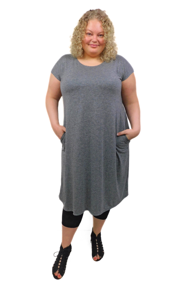 A woman plus size model wearing a gray long tunic dress with cap sleeves and side pockets, paired with black leggings and black strappy sandals.
