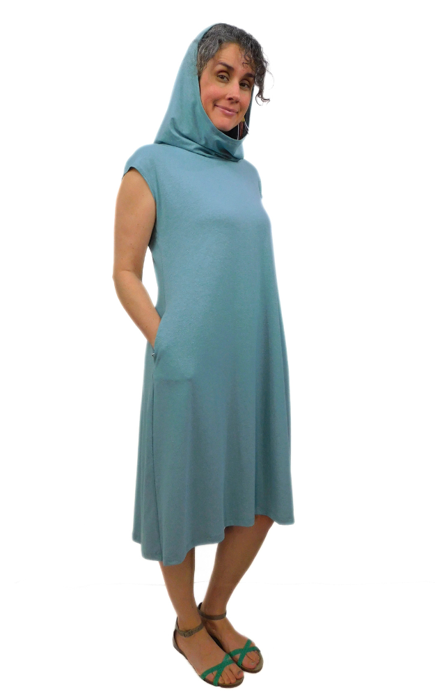 Woman standing with hands in pocket wearing light blue/green tunic with cap sleeves and large hood worn up.