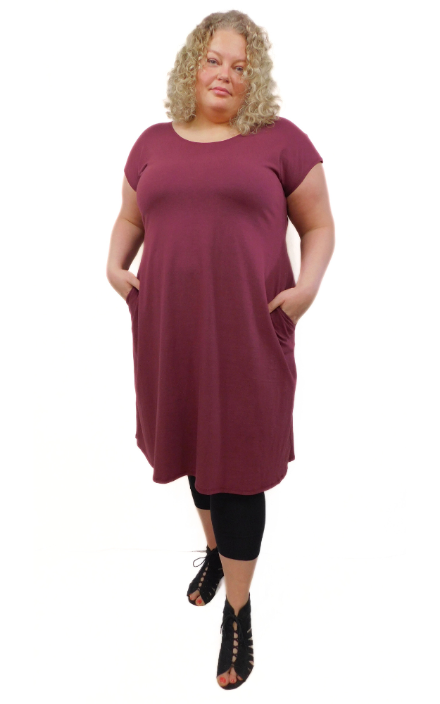 Plus size woman standing with hands in side pockets of long wine coloured tunic dress with cap sleeves and round neckline.