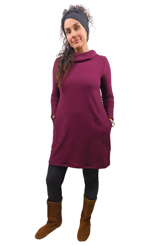 A woman modeling a long-sleeve cowl-neck tunic dress in deep burgundy, styled with a black headband, black leggings, and brown suede boots against a plain white background.