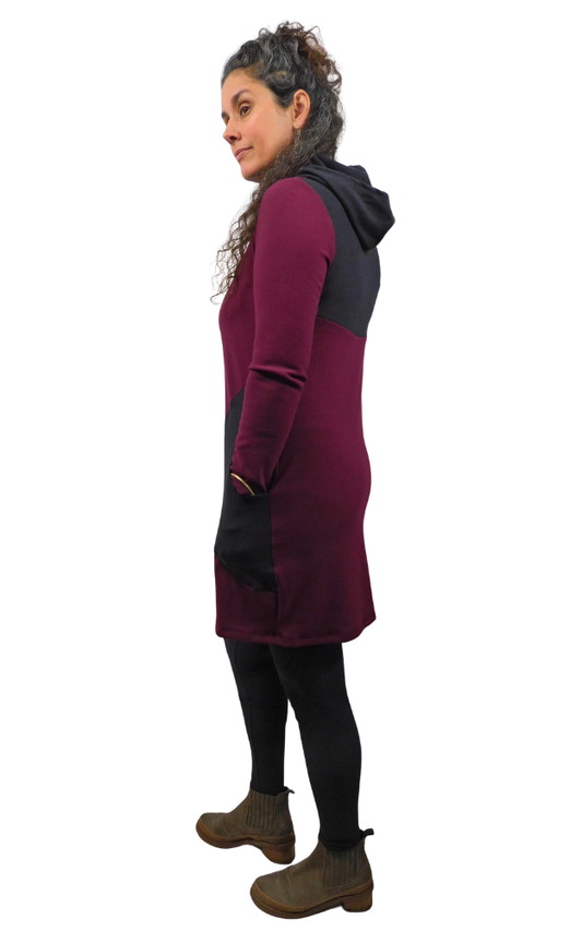 A woman modeling a color-blocked hooded tunic dress in burgundy and black, paired with black leggings and brown ankle boots, shown from a side profile against a plain white background.