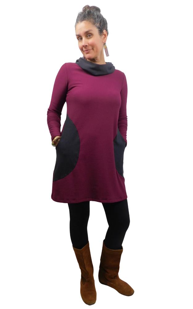 A woman modeling a long-sleeve cowl-neck (hood worn down) tunic dress in deep burgundy with contrasting dusty black panels on the front and pockets, styled with black leggings and brown boots against a plain white background.