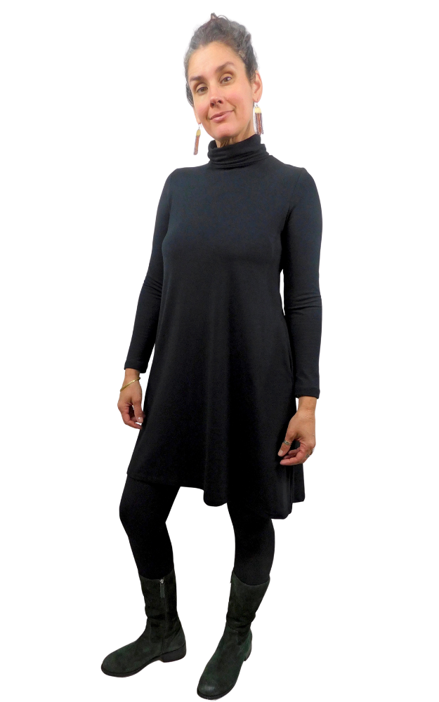 A woman modeling a black long-sleeve turtleneck dress with a flared silhouette, paired with black leggings and dark boots, against a plain white background.