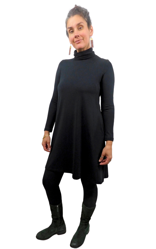 A woman modeling a black long-sleeve turtleneck dress with a flared silhouette, paired with black leggings and dark boots, against a plain white background.