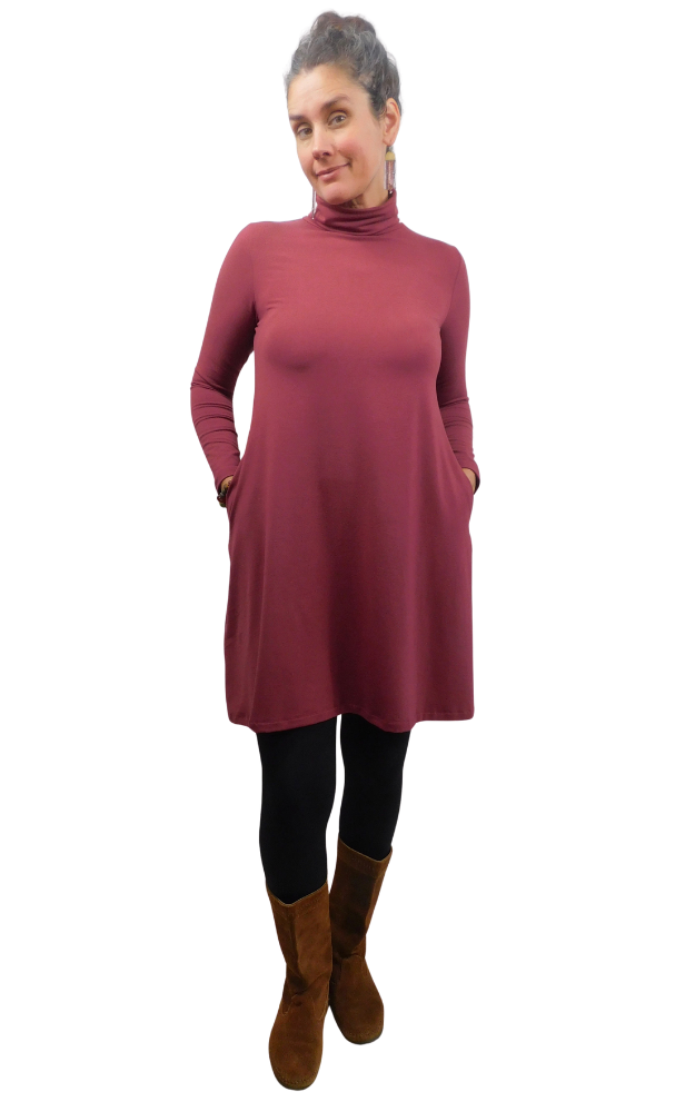 A woman modeling a long-sleeve, high-neck turtleneck dress in a dusty rose wine color, styled with black leggings and brown suede boots against a plain white background.
