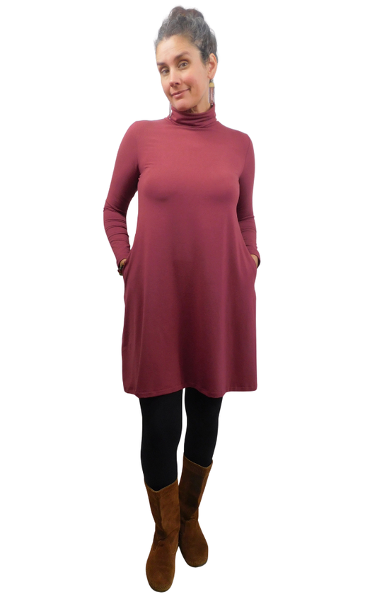 A woman modeling a long-sleeve, high-neck turtleneck dress in a dusty rose wine color, styled with black leggings and brown suede boots against a plain white background.