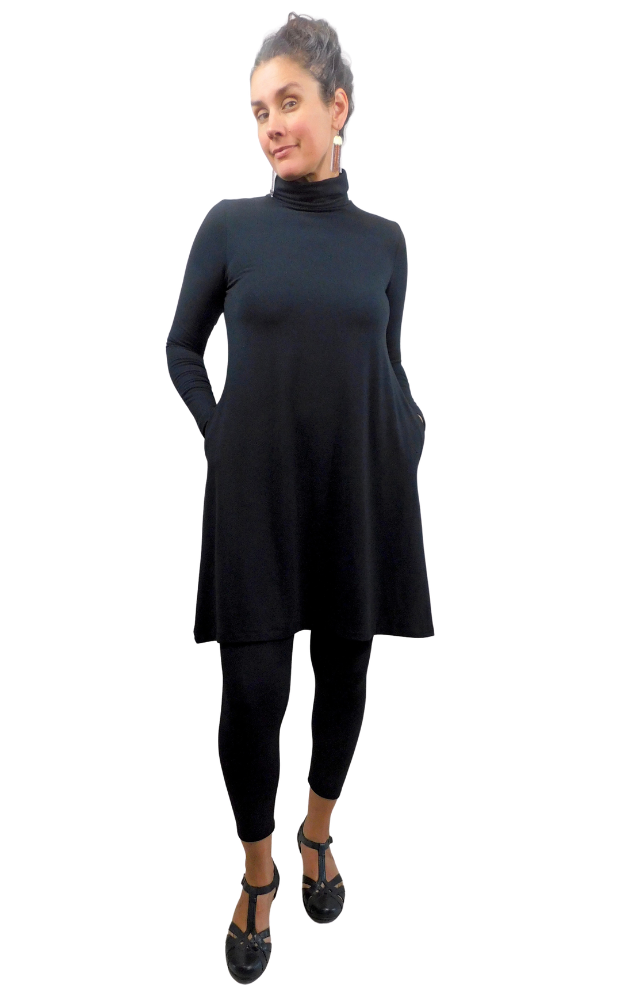 A woman modeling a black long-sleeve turtleneck dress with side pockets, worn over black leggings and black shoes, against a solid white background.
