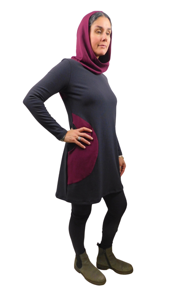 A woman modeling a long-sleeve tunic dress in black with side pockets and hood in deep burgandy, paired with black leggings and brown ankle boots, against a plain white background.