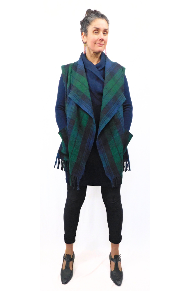 A woman wearing a green, blue, and black plaid  Merino Wool Tartan Vest Jacket with fringe, paired with a blue dress and black tights.