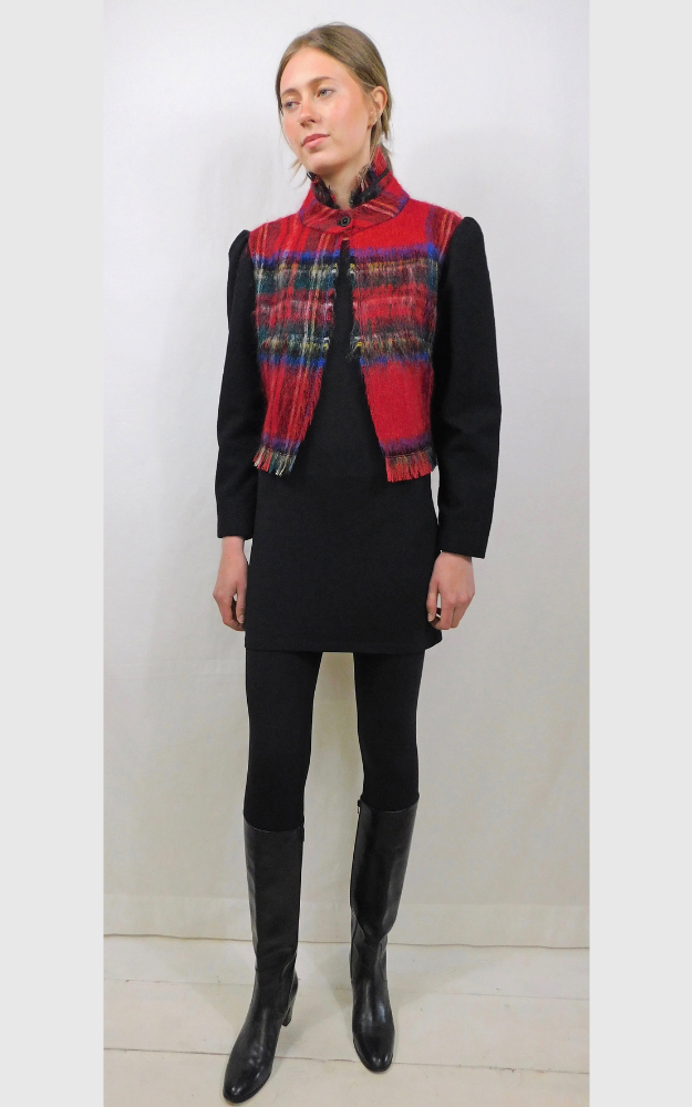 Woman wearing a red plaid jacket with collar and bottom fringe and plain long black sleeves on top of a black dress and black tights and black boots against a white background