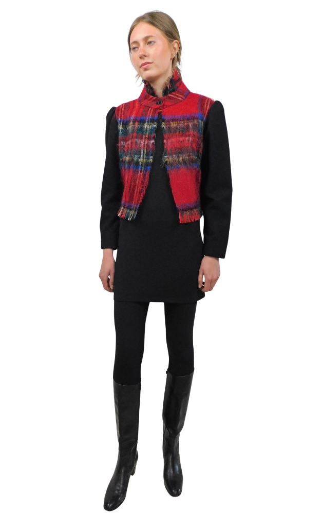 Woman wearing a red plaid jacket with collar and bottom fringe and plain long black sleeves on top of a black dress and black tights and black boots against a white background