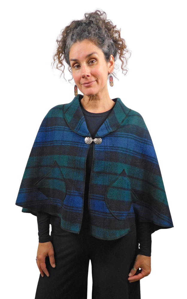 A woman modeling a blue and green plaid cape with a shawl collar, silver-tone button closure, and front patch pockets, photographed against a plain white background.