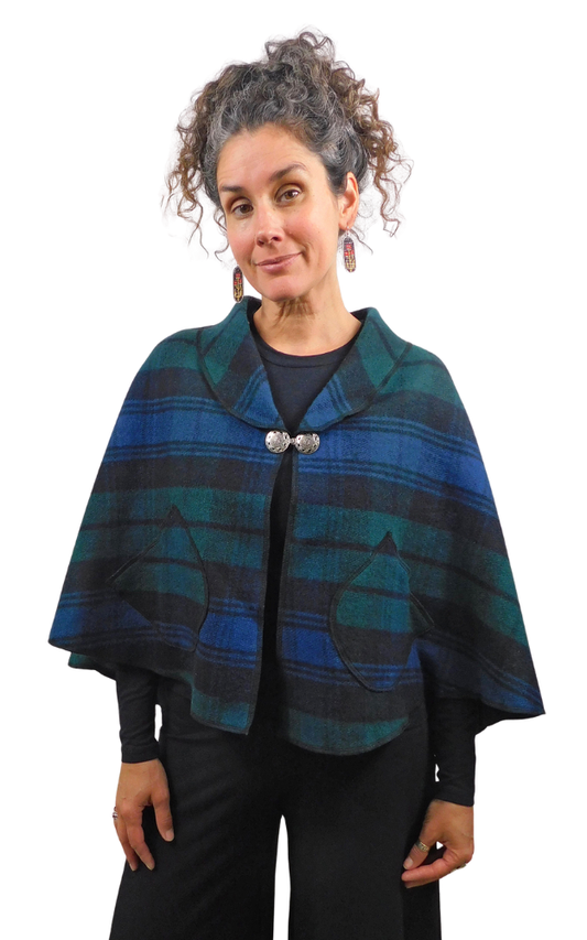 A woman modeling a blue and green plaid cape with a shawl collar, silver-tone button closure, and front patch pockets, photographed against a plain white background.