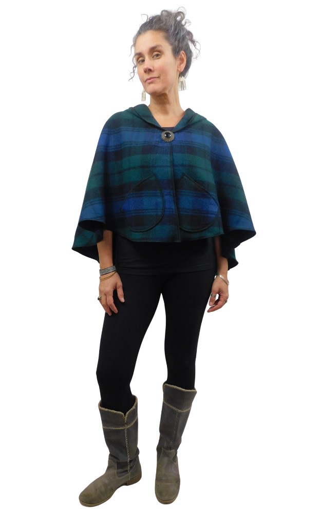 Woman wearing a blue and green and black tartan cape with front brooch and patch pockets worn over black leggings and grey boots 