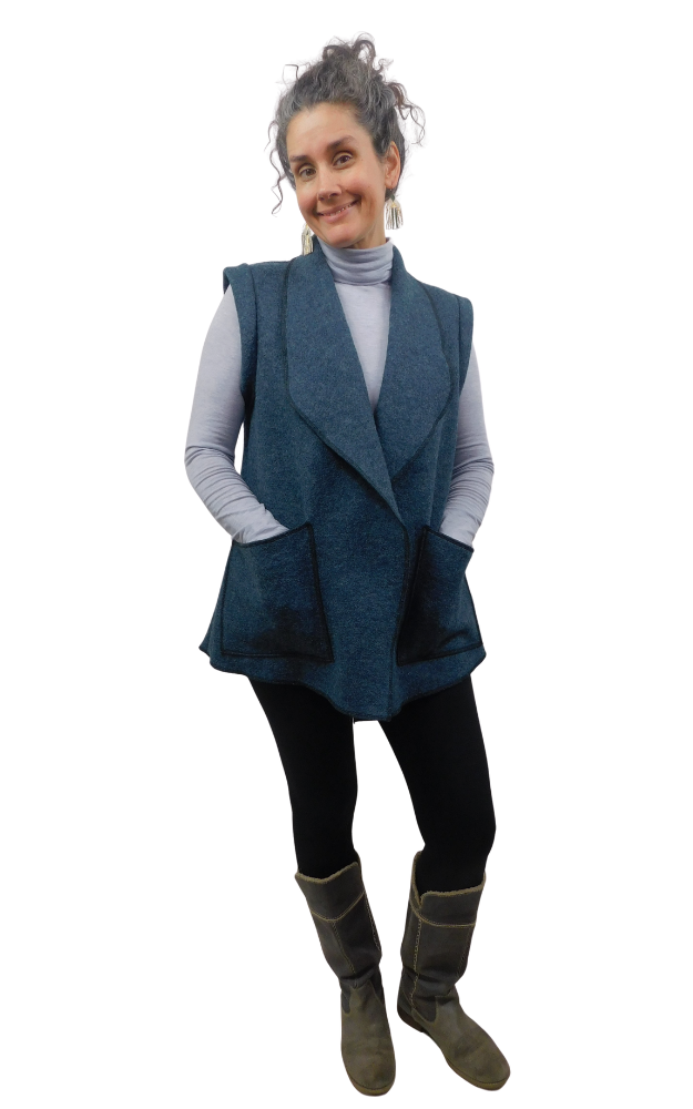 Woman wearing a dark blue heavy wool vest with large patch pockets, hands in pockets, wearing a long sleeve turtleneck grey sweater and black tights and grey boots
