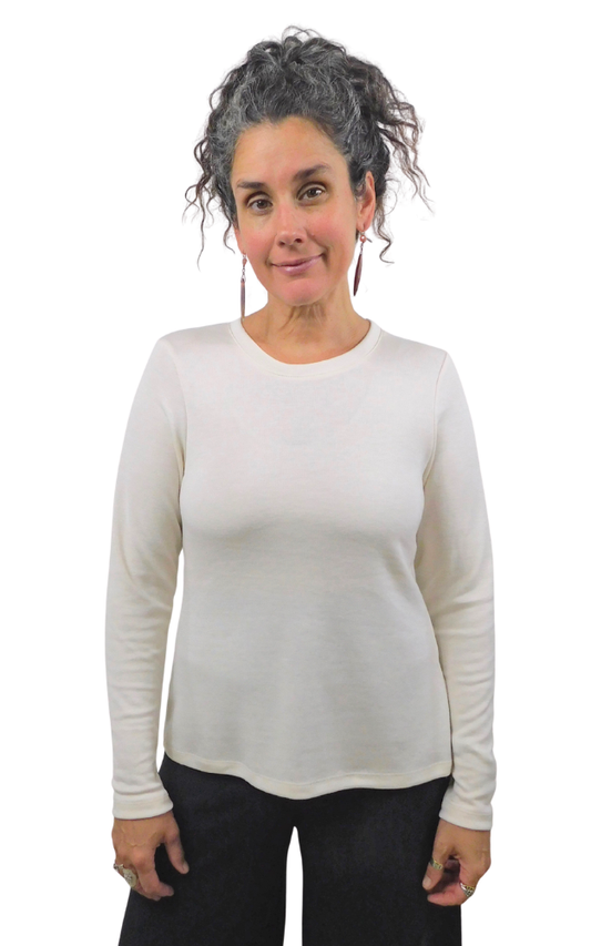 A woman with curly gray hair wearing a cream-colored long-sleeve crewneck top and black pants, standing against a solid white background.