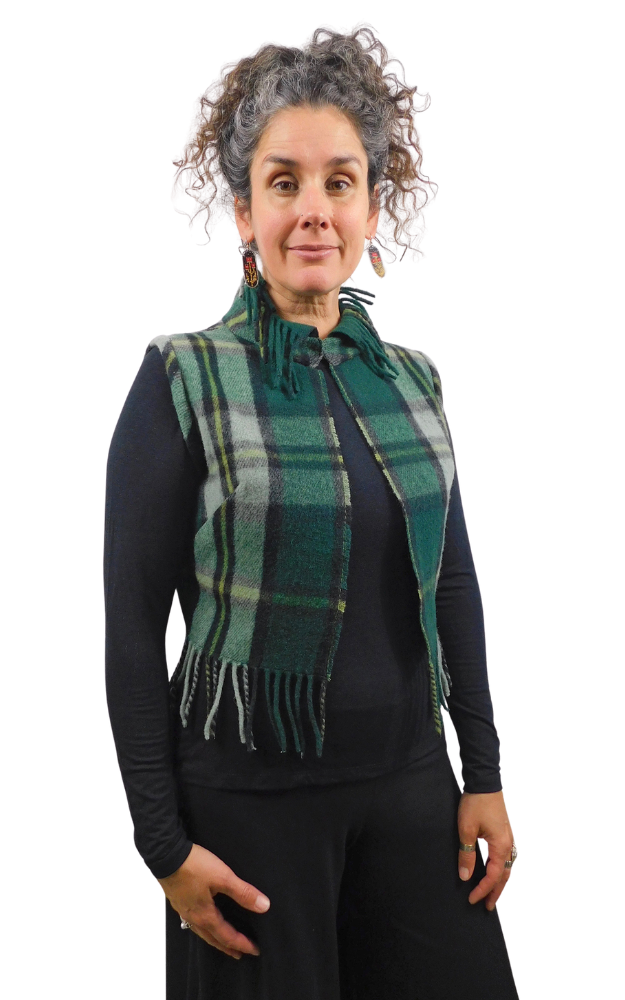 A woman with curly gray hair models a green and black plaid fringed vest over a black long-sleeve top, set against a plain white background.