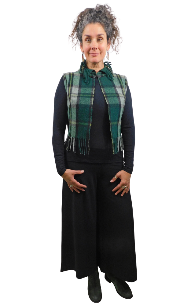 A woman modeling a green and black plaid fringed wrap vest over a black long-sleeve top and black wide-leg pants, standing against a solid white background.