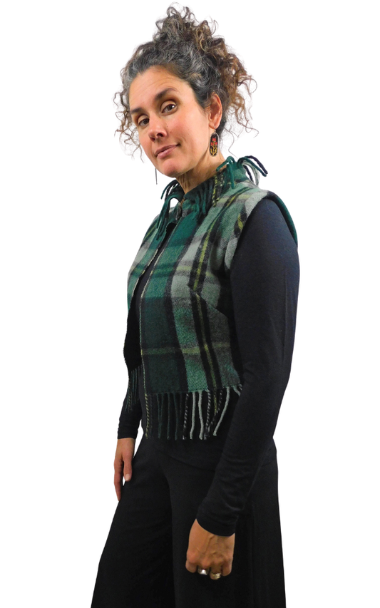 A woman wearing a green and black plaid fringed vest over a black long-sleeve top, photographed against a plain white background.