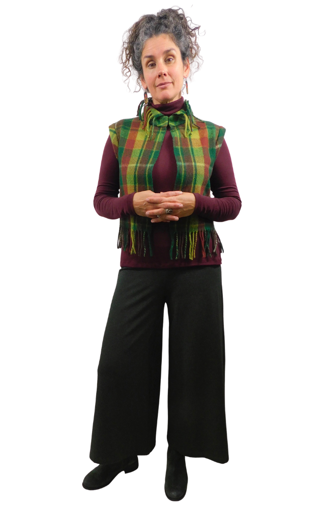 A woman modeling a green and yellow and wine tartan plaid fringed vest over a burgundy turtleneck, paired with black wide-leg pants and black shoes, against a plain white background.