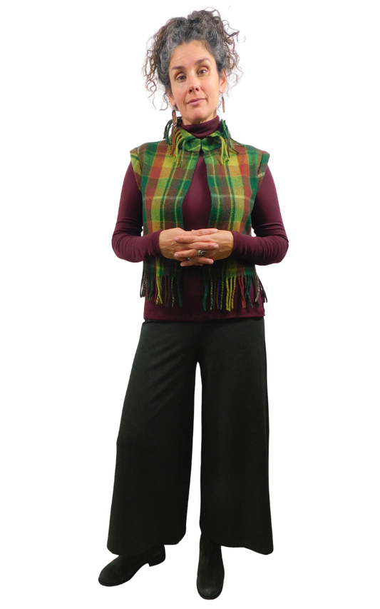 A woman modeling a green and yellow and wine tartan plaid fringed vest over a burgundy turtleneck, paired with black wide-leg pants and black shoes, against a plain white background.
