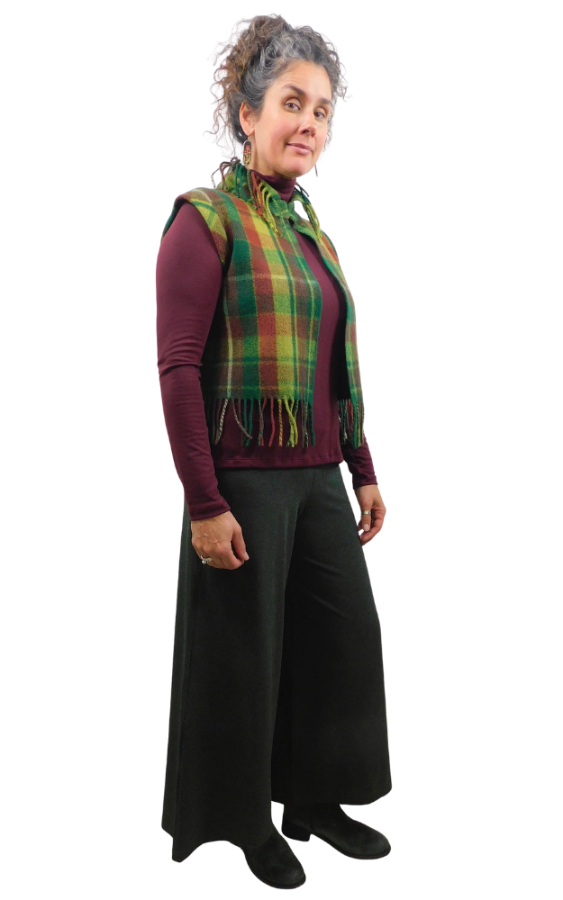 A woman modeling a green and yellow and wine tartan plaid fringed vest over a burgundy long-sleeve top and black wide-leg trousers, set against a plain white background.