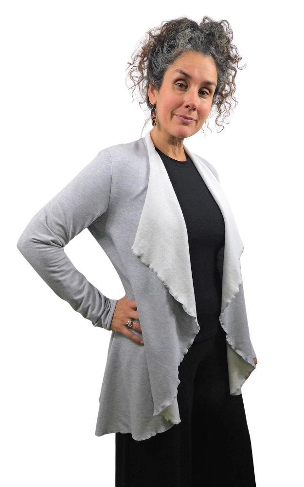 A woman with curly gray hair models a light gray long-sleeve open-front cardigan with a draped, frayed hem over a black top and black pants, set against a plain white background.