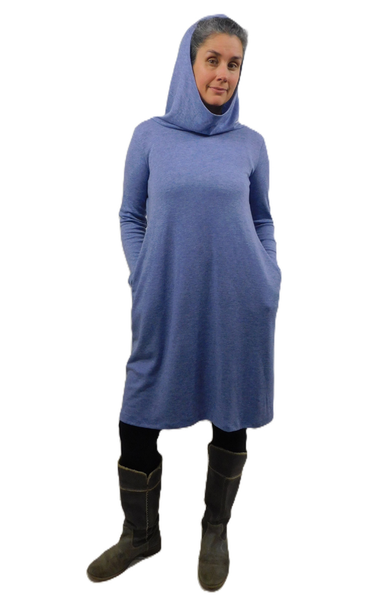 Woman wearing a long blue dress with long sleeves and hood with hands in pockets on top of black tights and grey boots