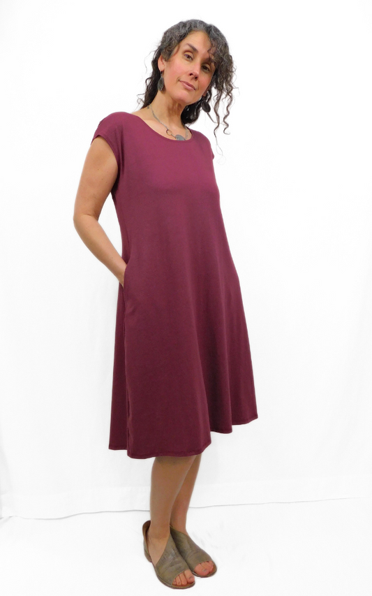 Woman standing with hands in side pockets of long wine coloured tunic dress with cap sleeves and round neckline.