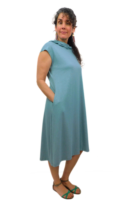 Woman standing with hands in pocket wearing  light blue/green tunic with cap sleeves and large hood worn down.