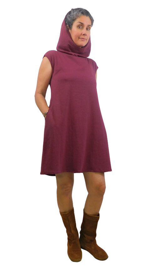 Woman wearing a maroon sleeveless dress with hood and hands in pocket and brown boots on a white background