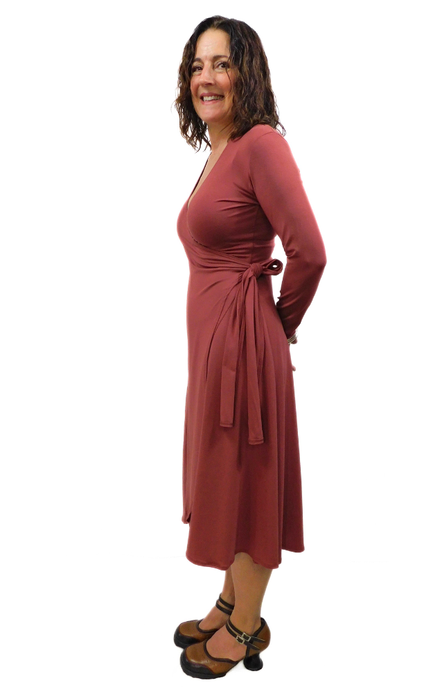 Woman standing sideways hands behind back wearing wine coloured wrap dress long sleeves tie belt below knee hem