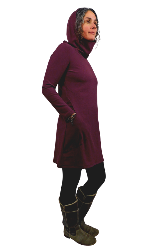 A woman modeling a long-sleeve, high-neck hooded tunic dress in deep burgundy, paired with black leggings and brown boots, shown against a plain white background.