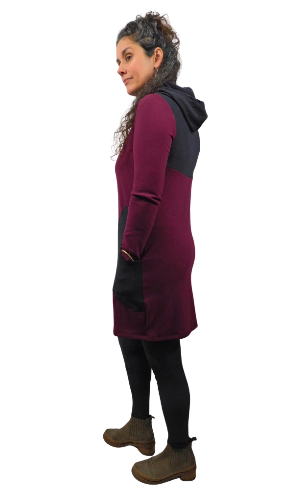 A woman modeling a color-blocked hooded tunic dress in burgundy and black, paired with black leggings and brown ankle boots, shown from a side profile against a plain white background.