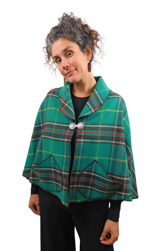 A woman modeling a green tartan plaid cape poncho with silver button closures, worn over a black top and black pants, against a plain white background.