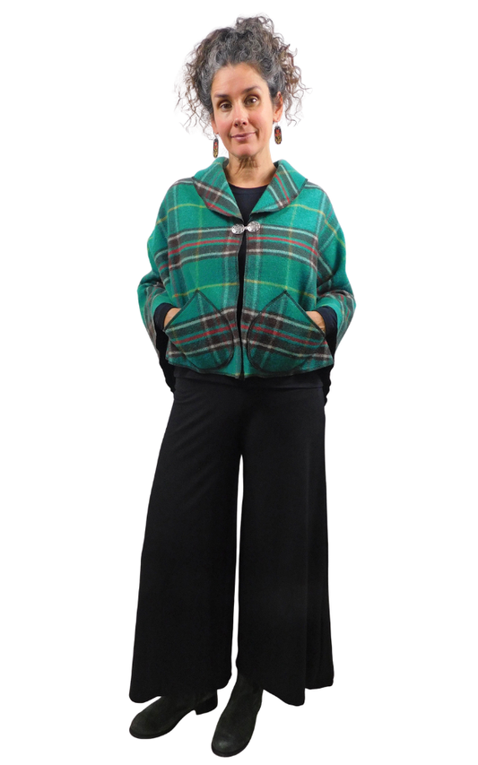 A woman modeling a green tartan plaid cape with front pockets, paired with black wide-leg pants and black boots, against a plain white background.