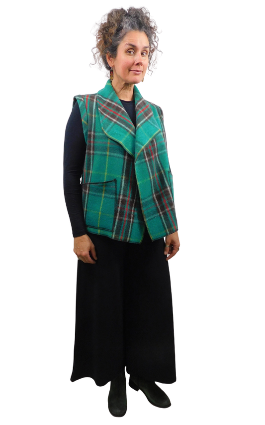 A woman modeling a sleeveless green tartan plaid vest over a black long-sleeve top and black wide-leg trousers, photographed against a plain white background.