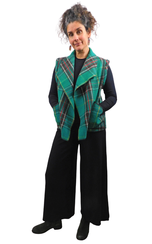 A woman modeling a sleeveless green and black tartan plaid vest over a black top and wide-leg black pants, photographed against a plain white background.