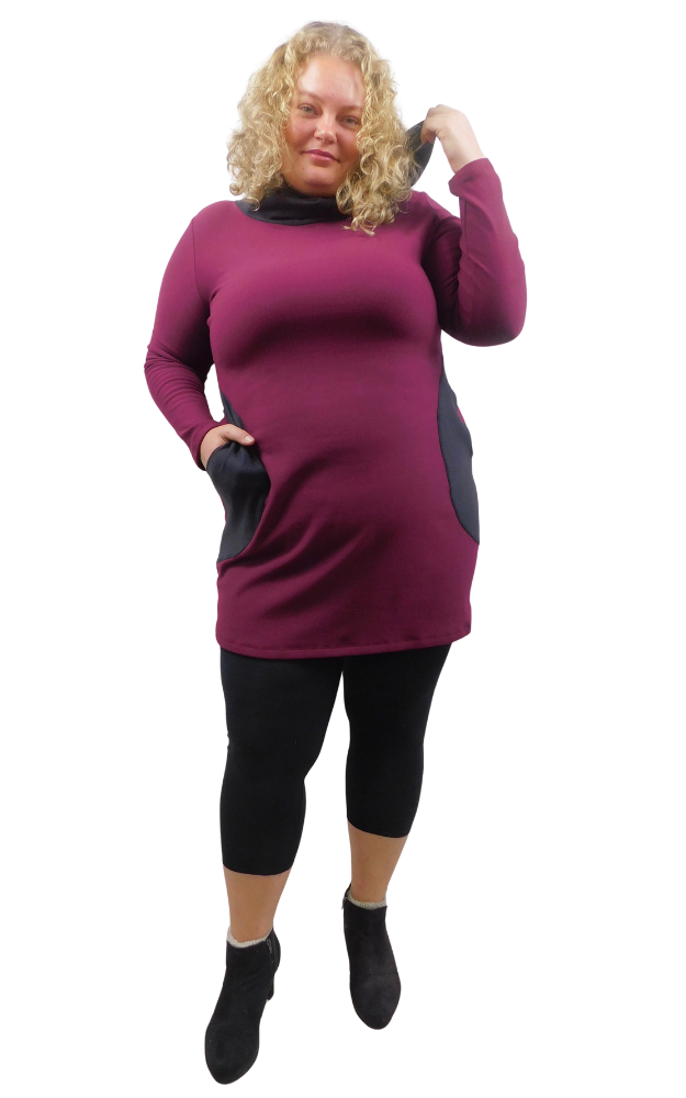 A woman modeling a long-sleeve cowl-neck (hood worn down) tunic dress in deep burgundy with contrasting dusty black panels on the front pockets, styled with black leggings and brown boots against a plain white background.