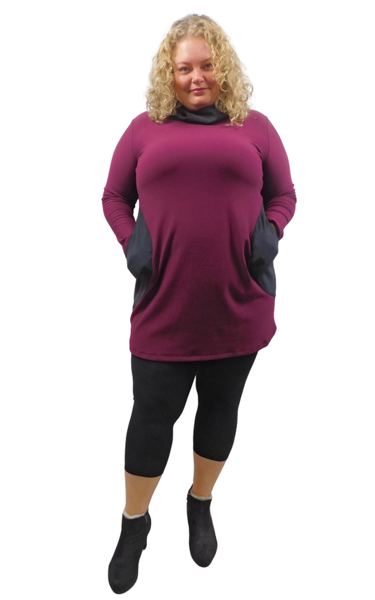 A woman modeling a long-sleeve cowl-neck (hood worn down) tunic dress in deep burgundy with contrasting dusty black front pockets, styled with black leggings and brown boots against a plain white background.