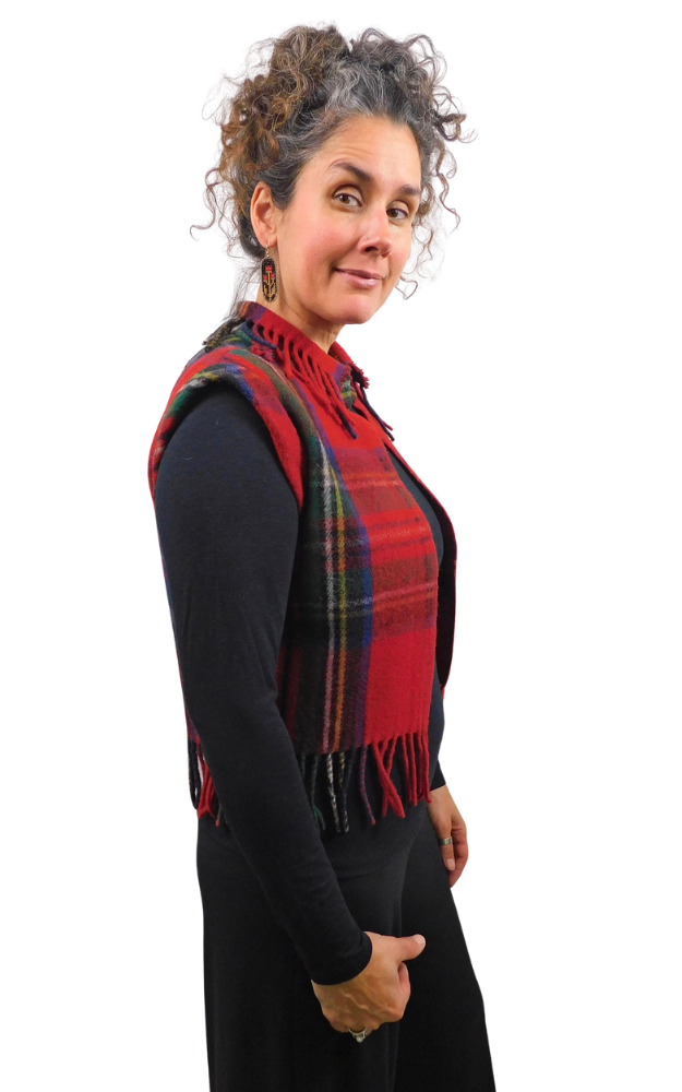 A woman with curly gray hair is shown from the side, wearing a red and black tartan plaid fringed vest over a black long-sleeve top, against a solid white background.