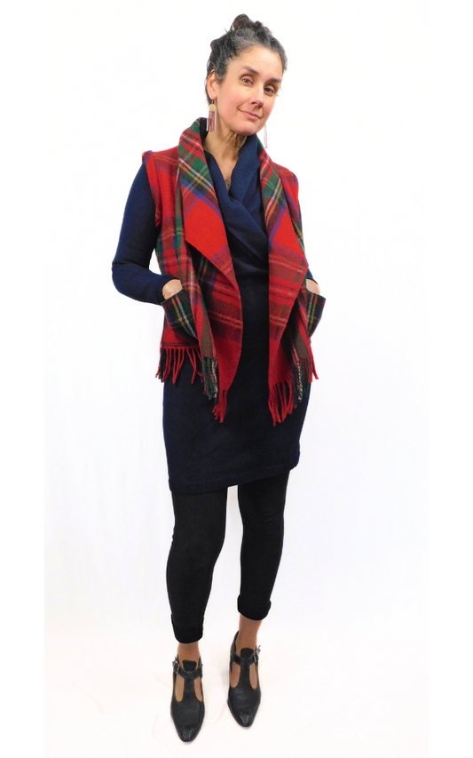 A woman wearing a black and red tartan patterned vest with fringe detail, paired with black trousers and black shoes, standing against a white background.